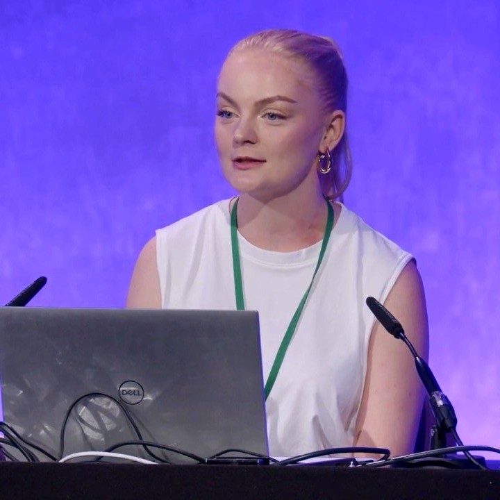 a blond haired white woman at a podium, ready to speak, in a while shirt with a purple backdrop.